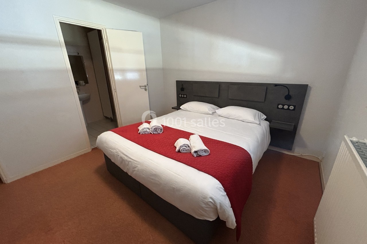 Chambre avec lit double, draps blancs, couverture rouge, serviettes pliées et accès à une salle de bain attenante.