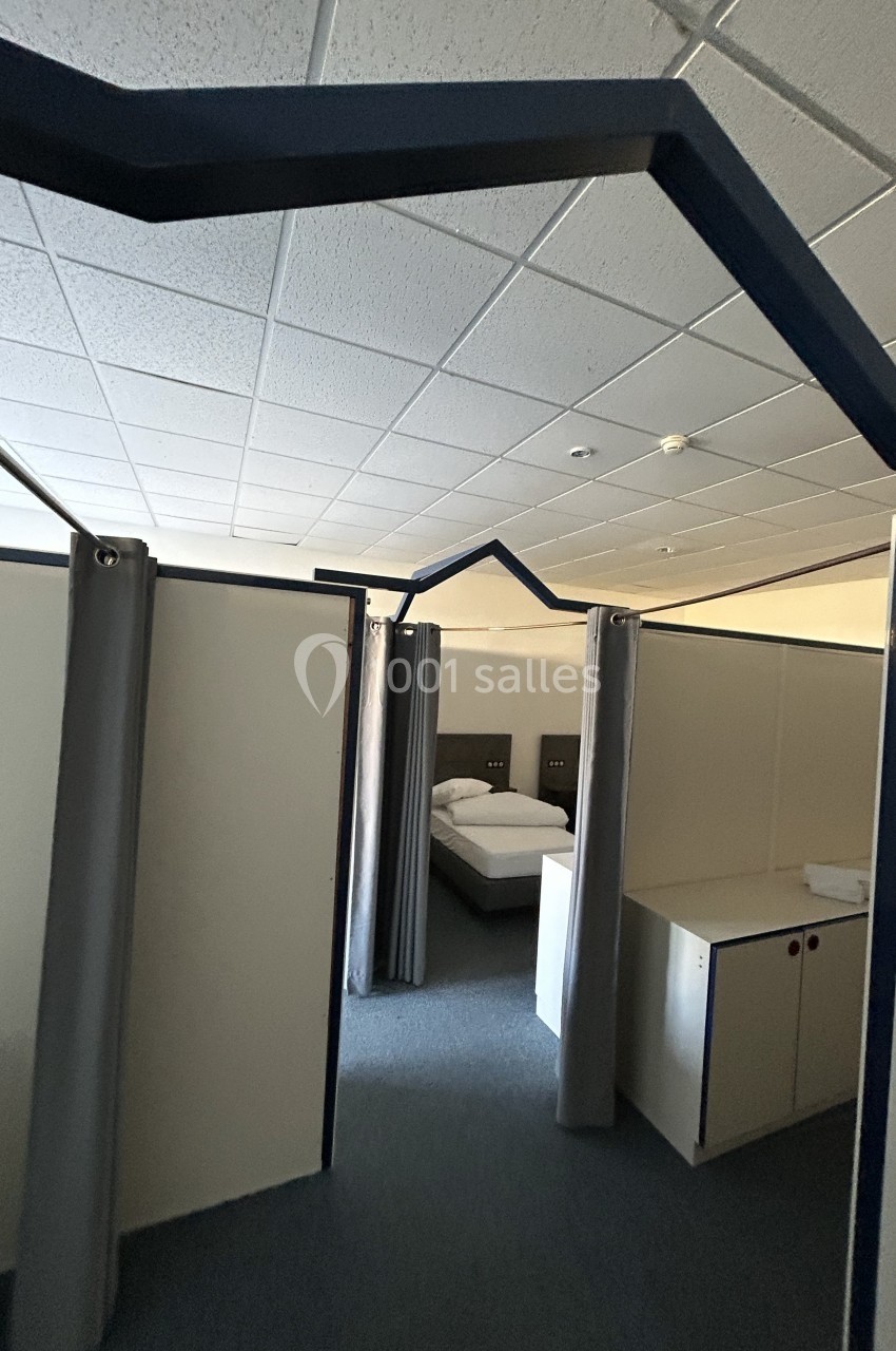 Cloisons modulaires avec rideaux séparant des espaces, dont un contenant un lit et un meuble, dans une pièce éclairée.