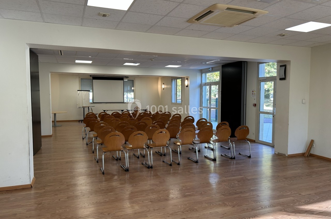 Salle de conférence lumineuse avec des rangées de chaises en bois, un écran de projection et un tableau blanc.