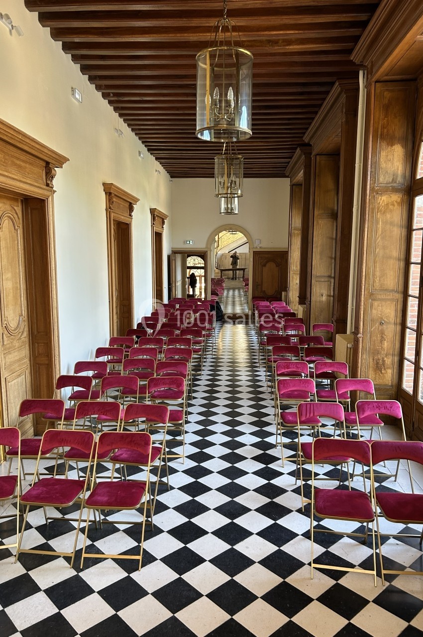 Rangées de chaises pliantes rouges alignées dans un couloir avec sol en damier et boiseries.