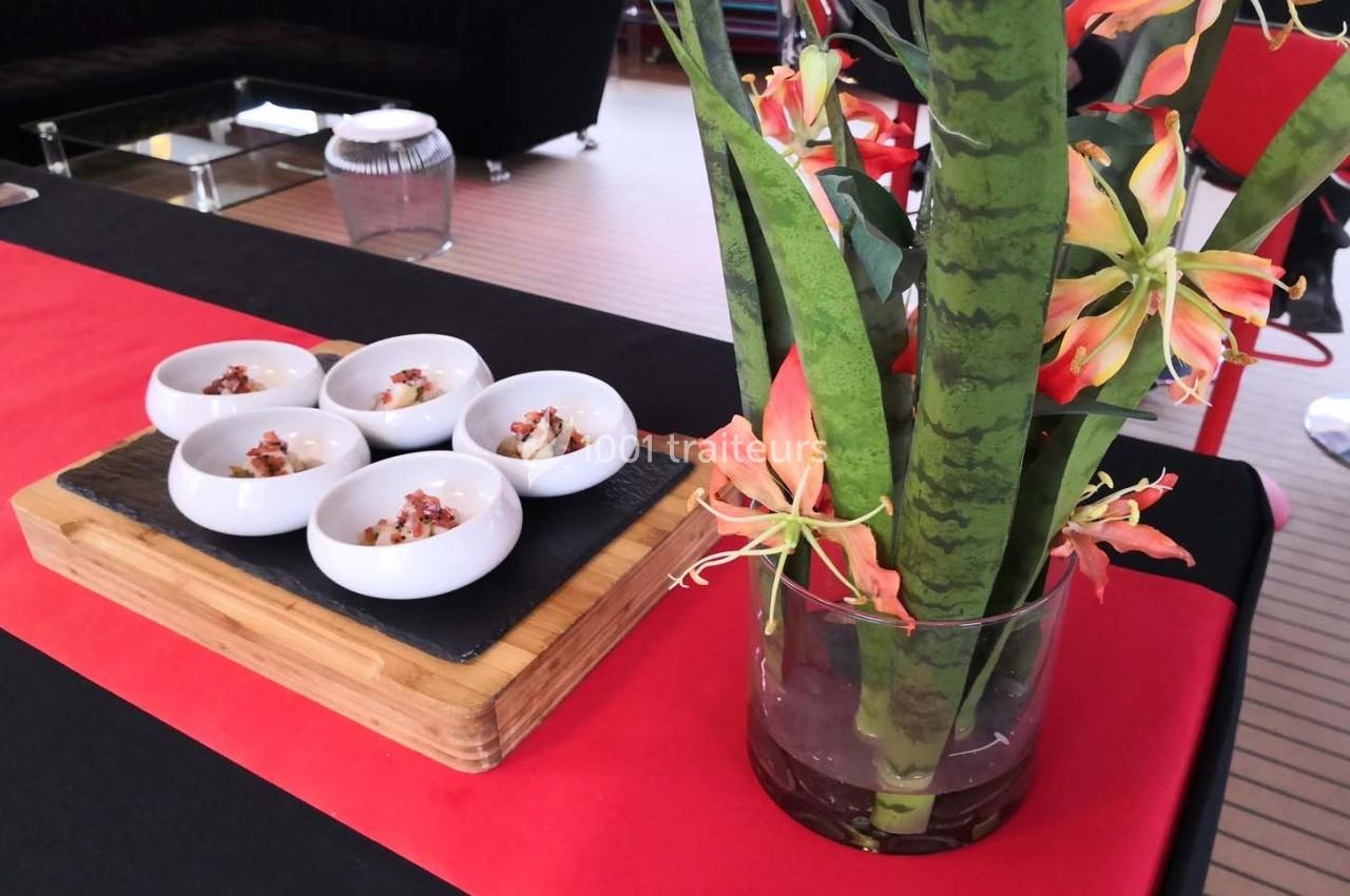 Six petites coupelles blanches contenant des amuse-bouches, disposées sur une planche en bois près d'un vase fleuri.