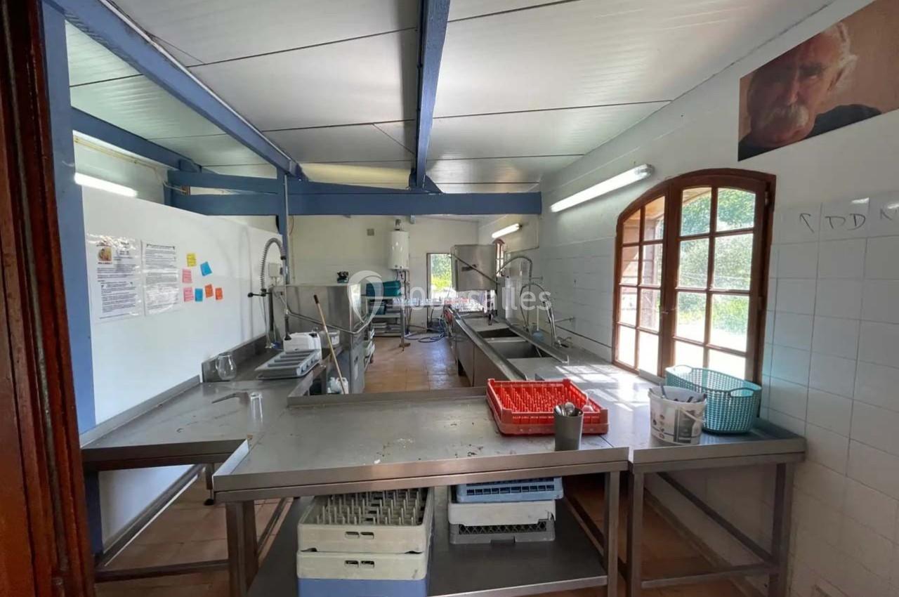 Cuisine collective avec plans de travail en inox, éviers, paniers de vaisselle et lumière naturelle entrant par une fenêtre.