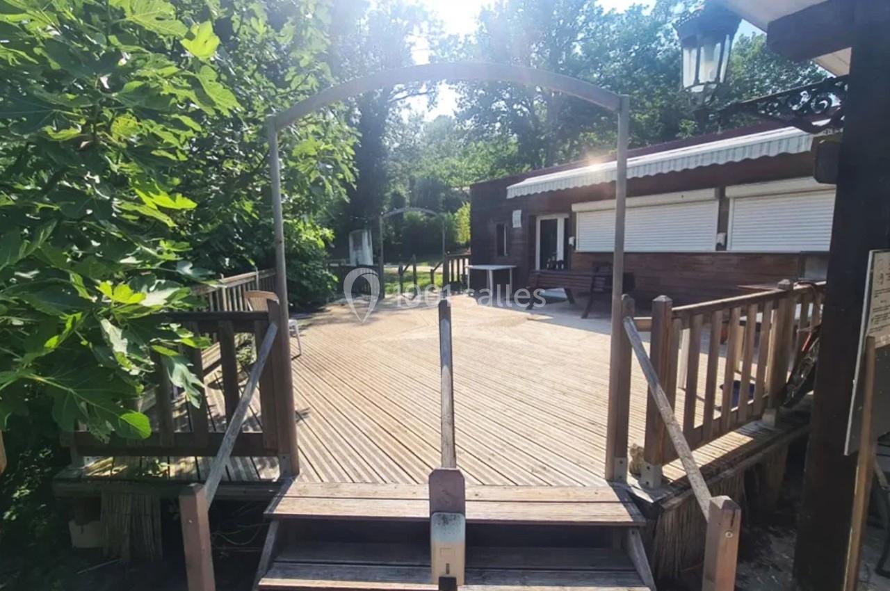 Passerelle en bois menant à une terrasse entourée de végétation, avec un bâtiment en arrière-plan.