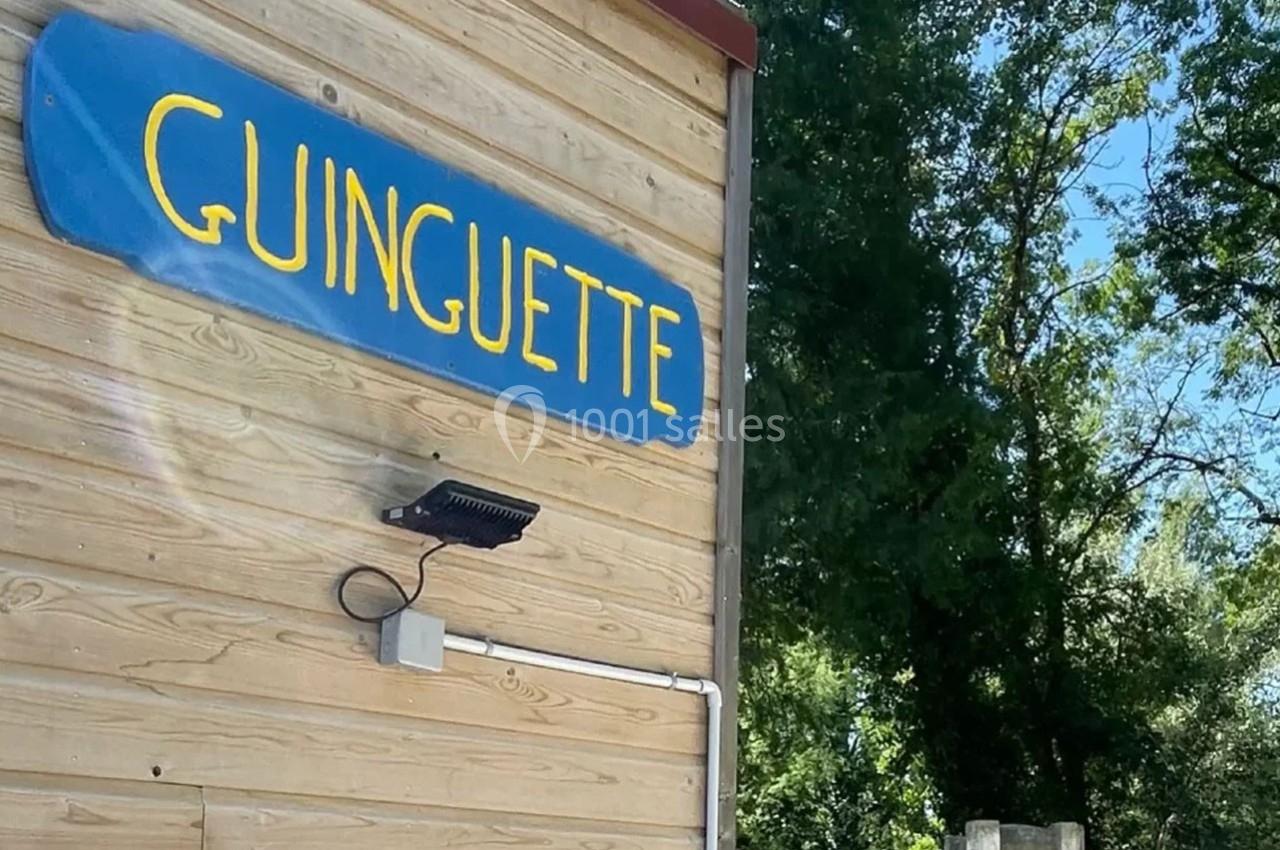 Panneau bleu marqué ’Guinguette’ fixé sur un mur en bois, entouré de verdure en arrière-plan.