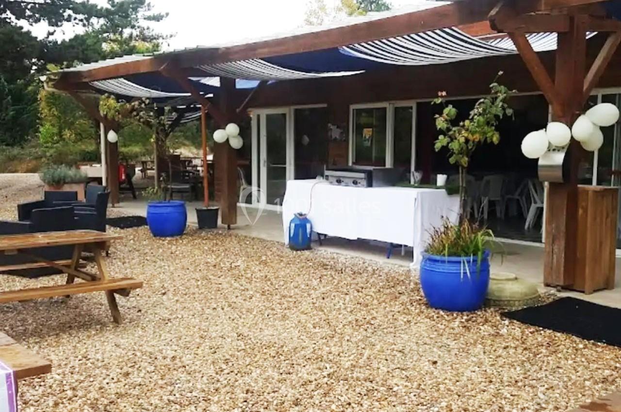Terrasse extérieure avec tables en bois, pots bleus, décorations blanches et store rayé, devant un bâtiment vitré.