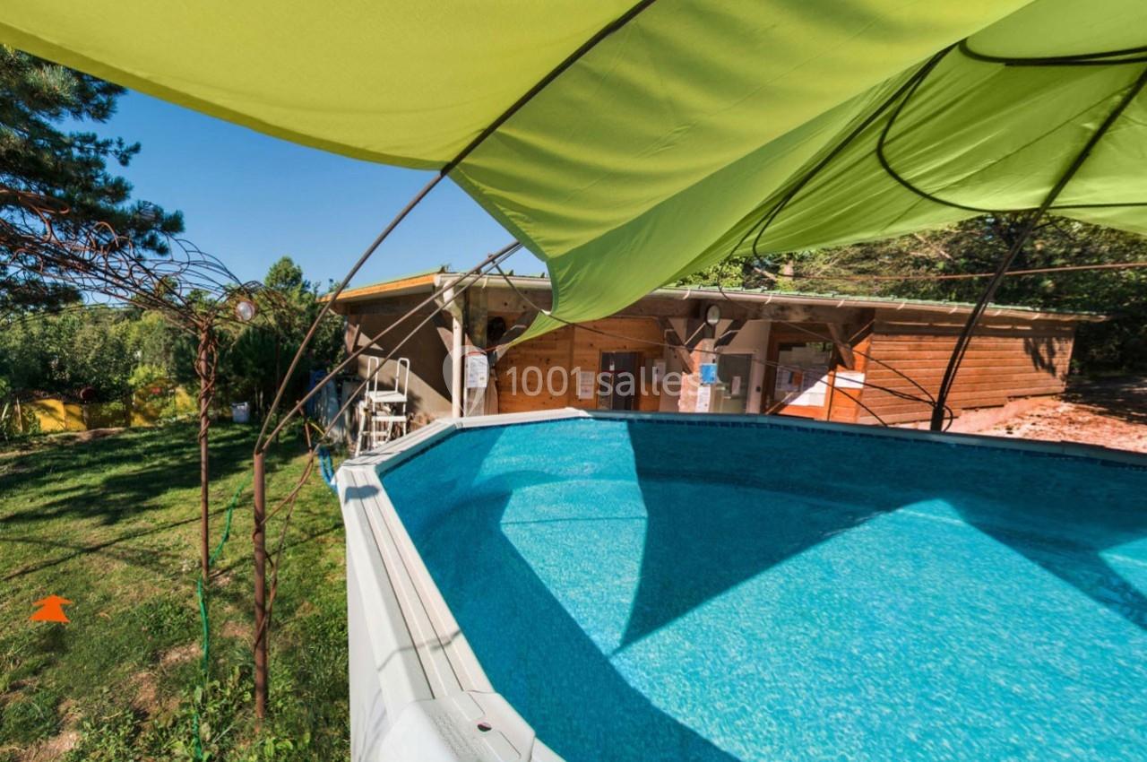 Piscine hors-sol entourée de verdure avec un abri en bois en arrière-plan, vue partielle d'un auvent vert.