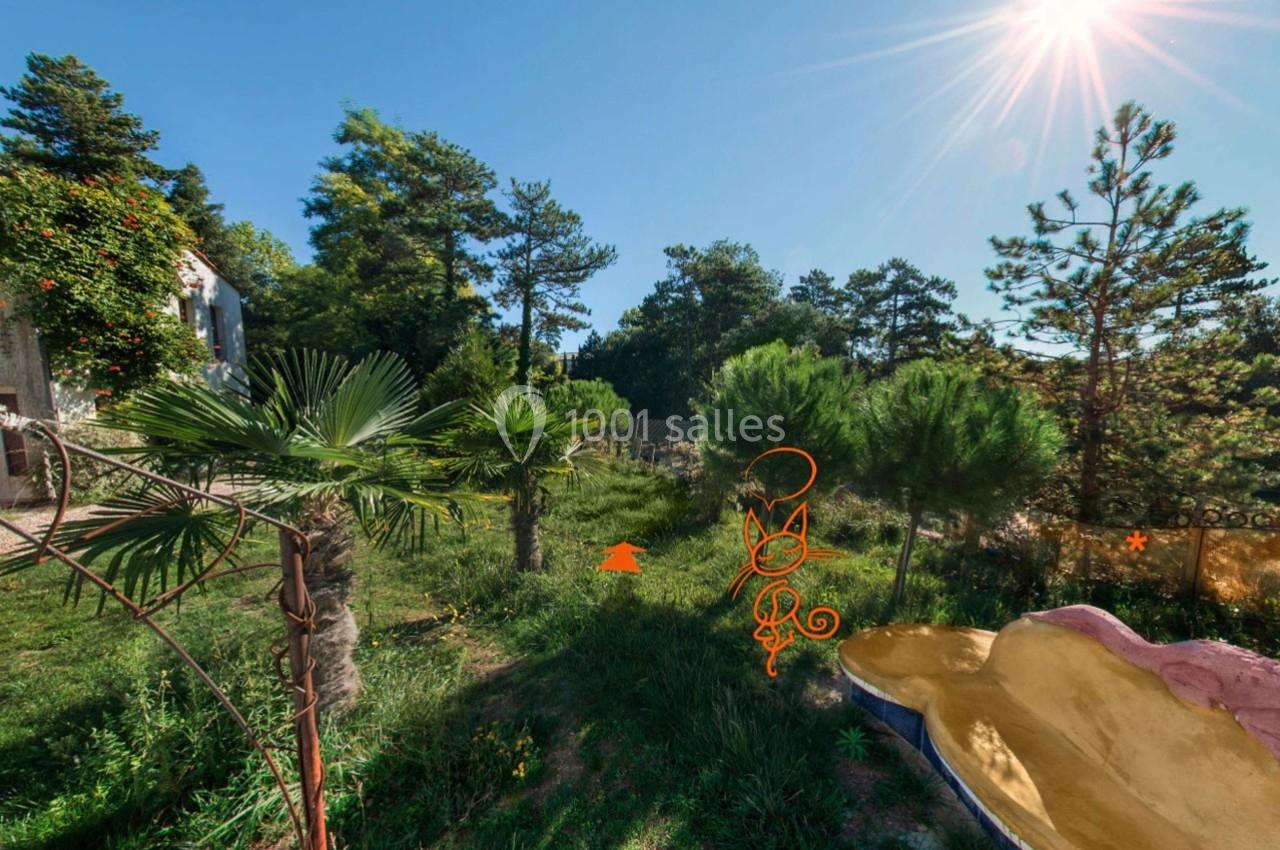 Jardin verdoyant avec des palmiers, des pins et des buissons, baigné de soleil sous un ciel bleu clair.