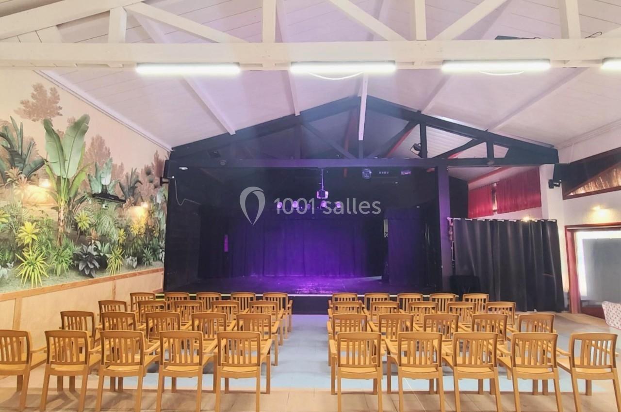 Salle avec des chaises en bois alignées face à une scène éclairée de lumières violettes, décor mural végétal.