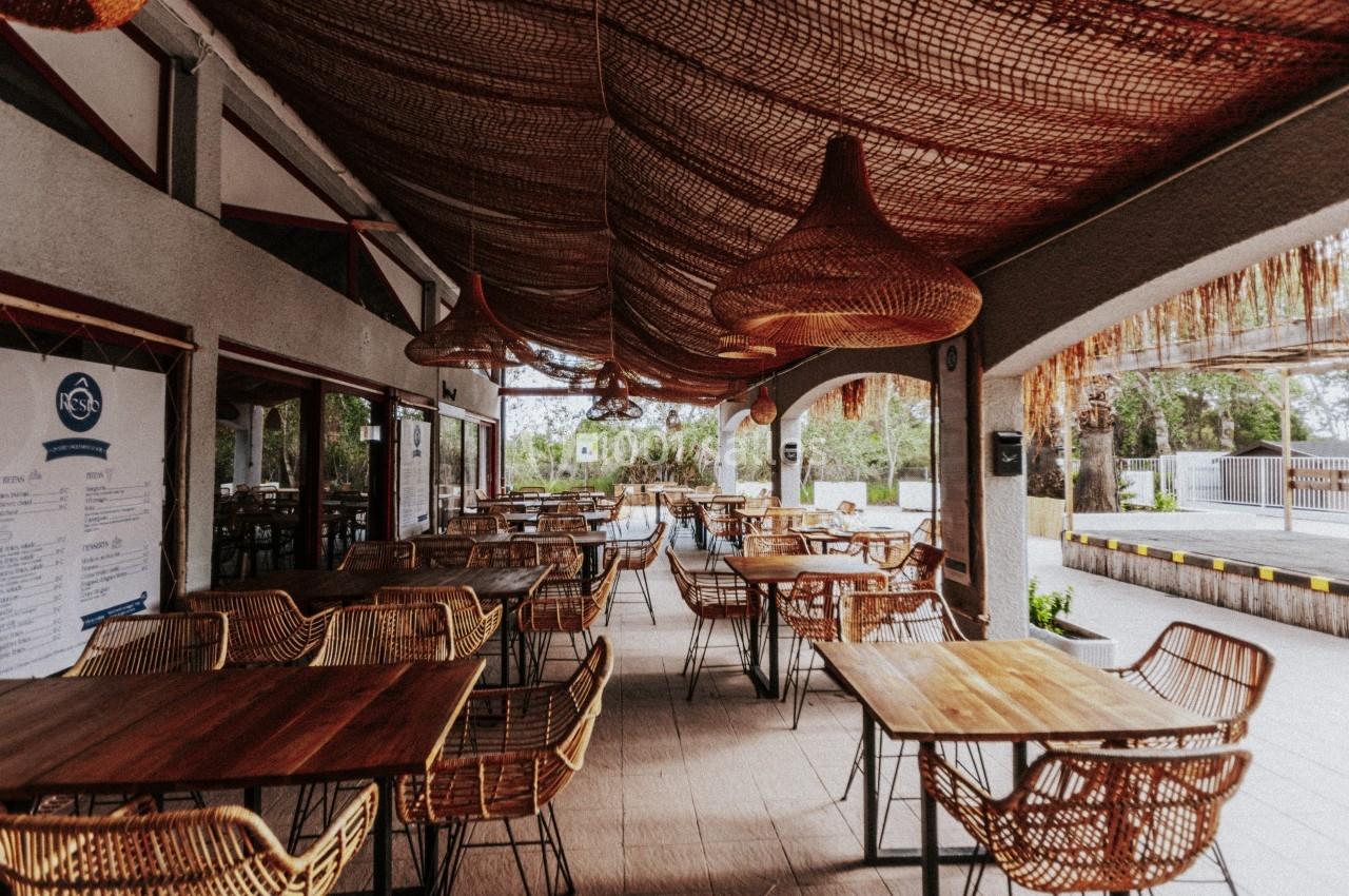 Terrasse couverte avec tables en bois et chaises en osier, décorée de suspensions en rotin, donnant sur un espace extérieur.