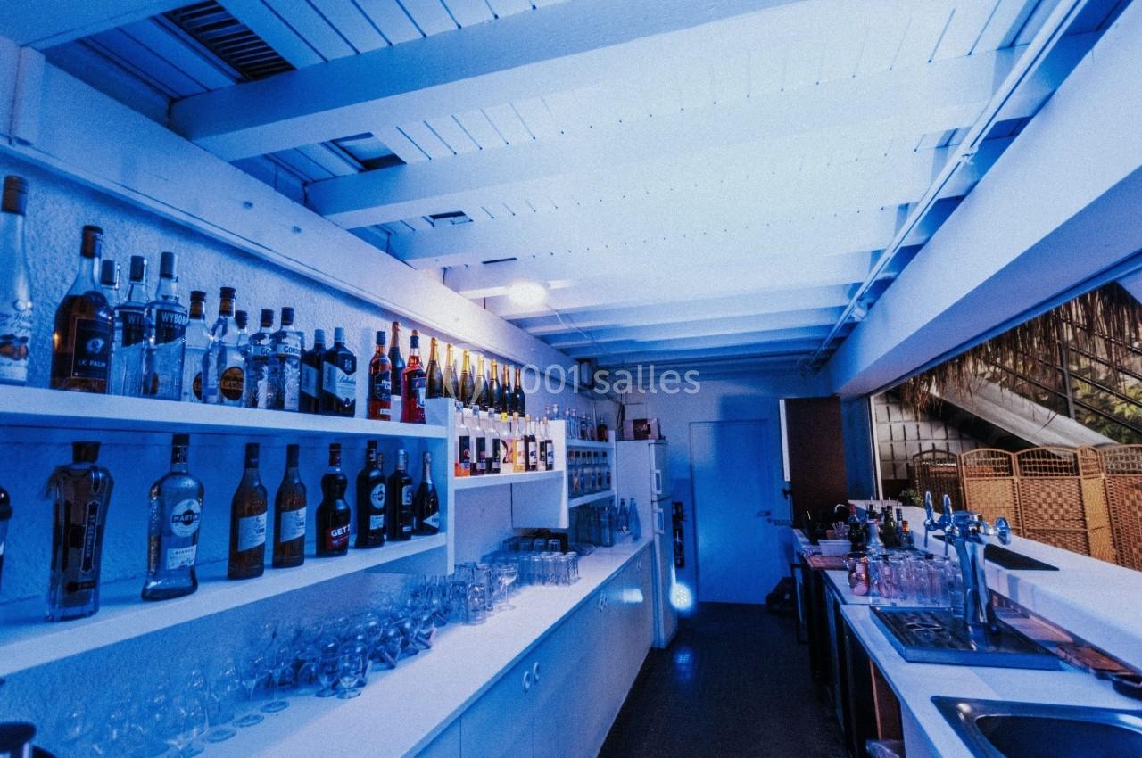 Intérieur d'un bar moderne avec étagères remplies de bouteilles et un comptoir éclairé par une lumière bleue.