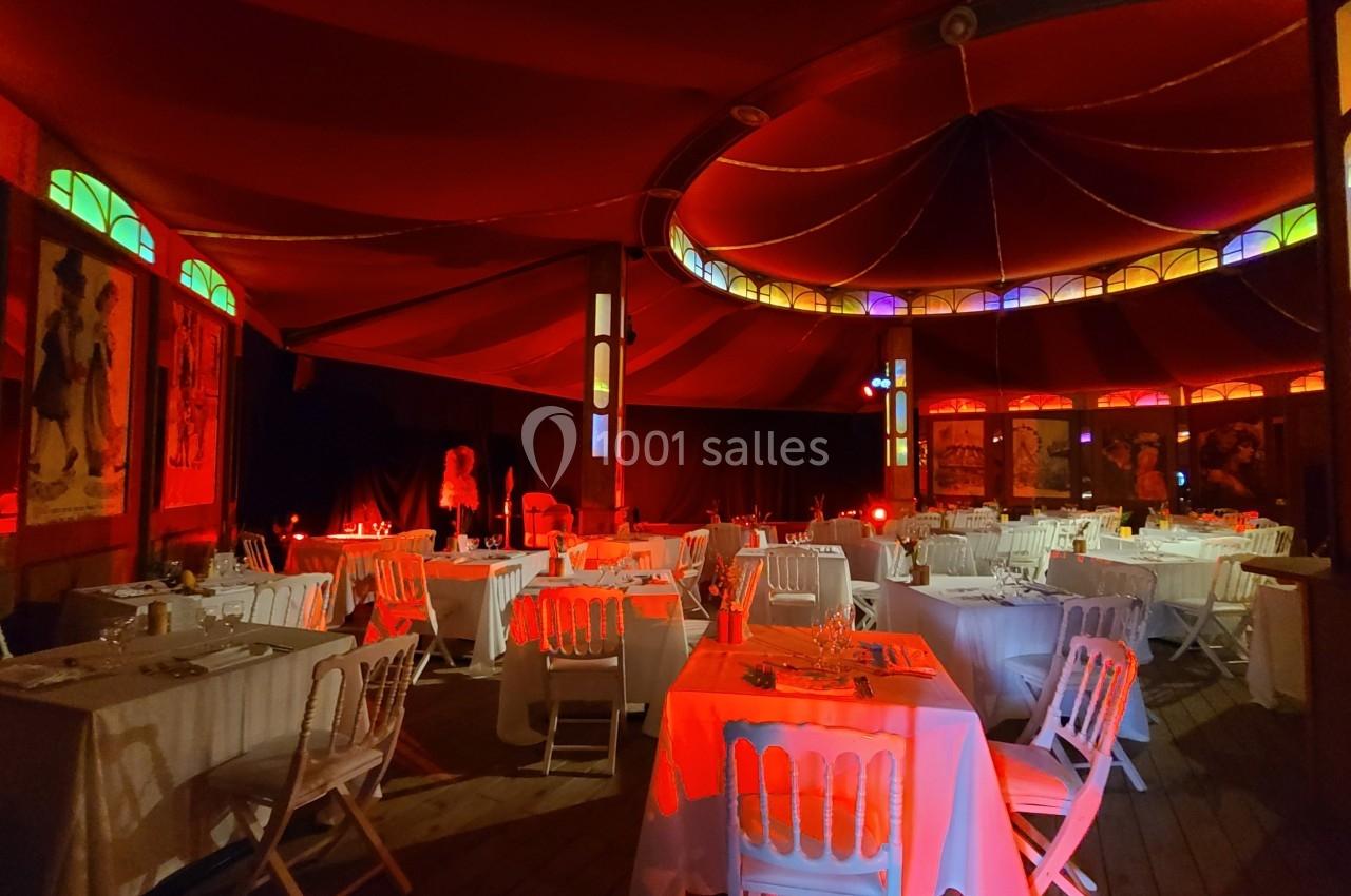 Salle de réception sous un chapiteau rouge avec tables dressées et éclairage tamisé coloré.