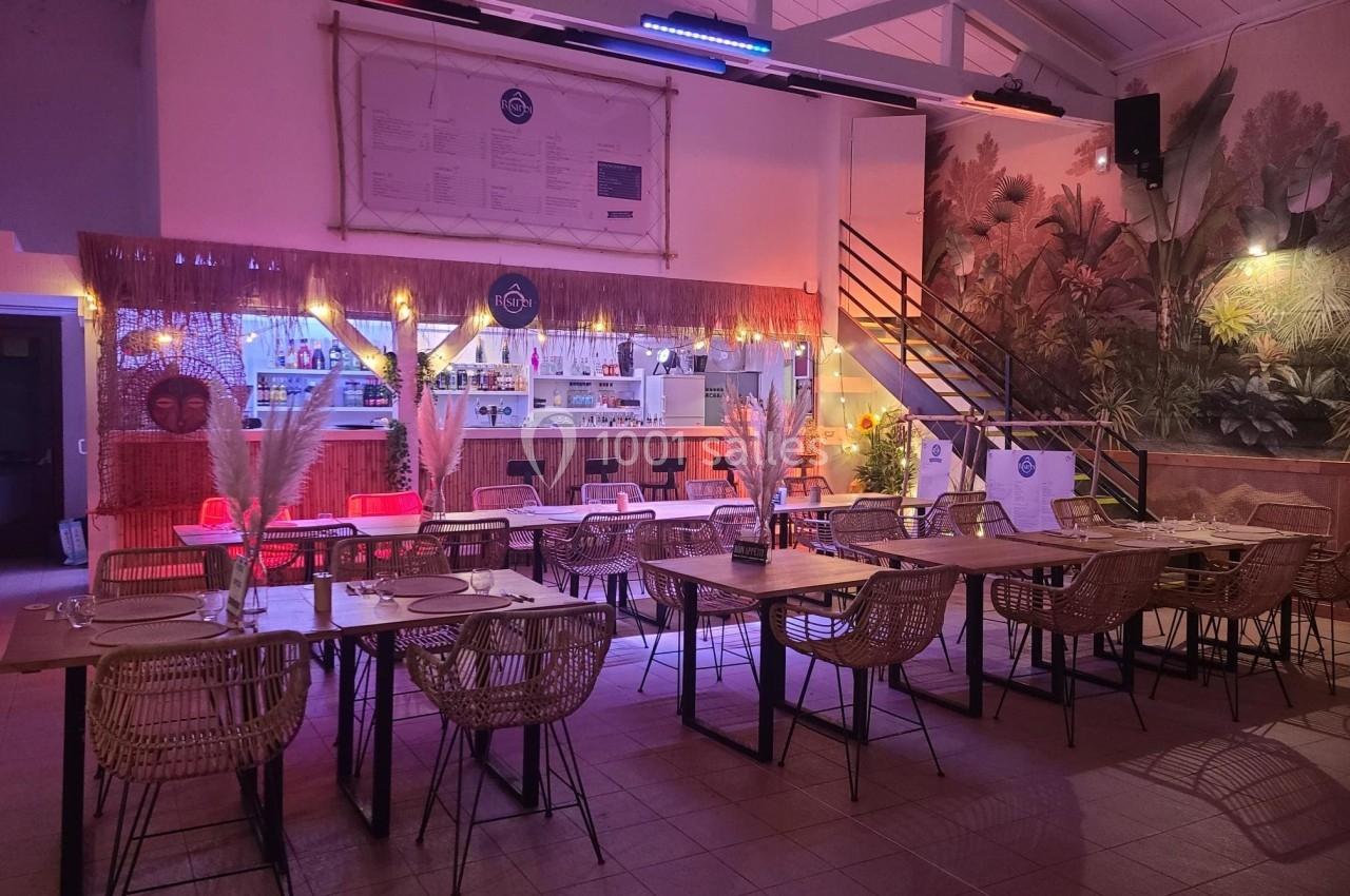 Salle de restaurant décorée avec des tables en bois, des chaises en rotin et un éclairage tamisé coloré.