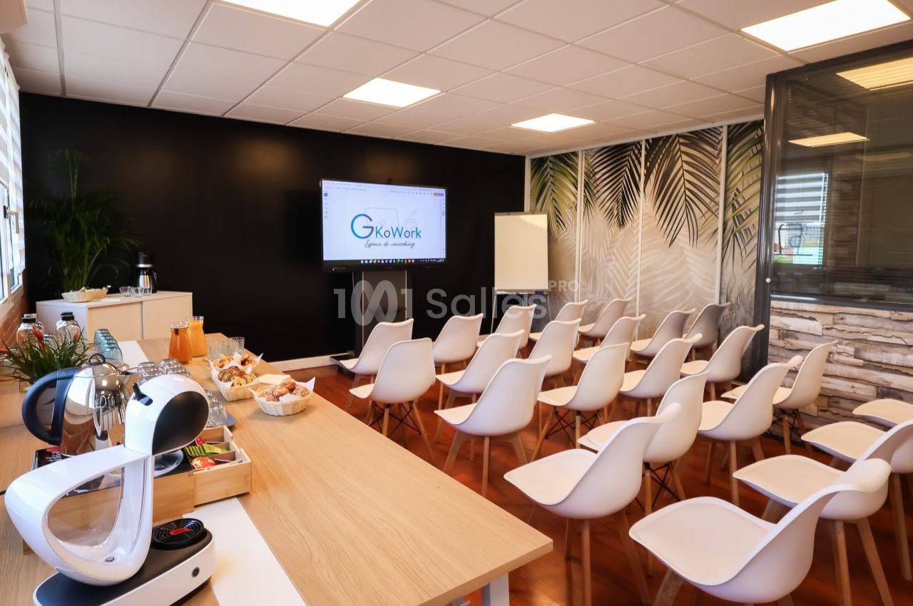 Salle de réunion avec chaises blanches alignées, écran de présentation, table avec collations et décoration végétale.