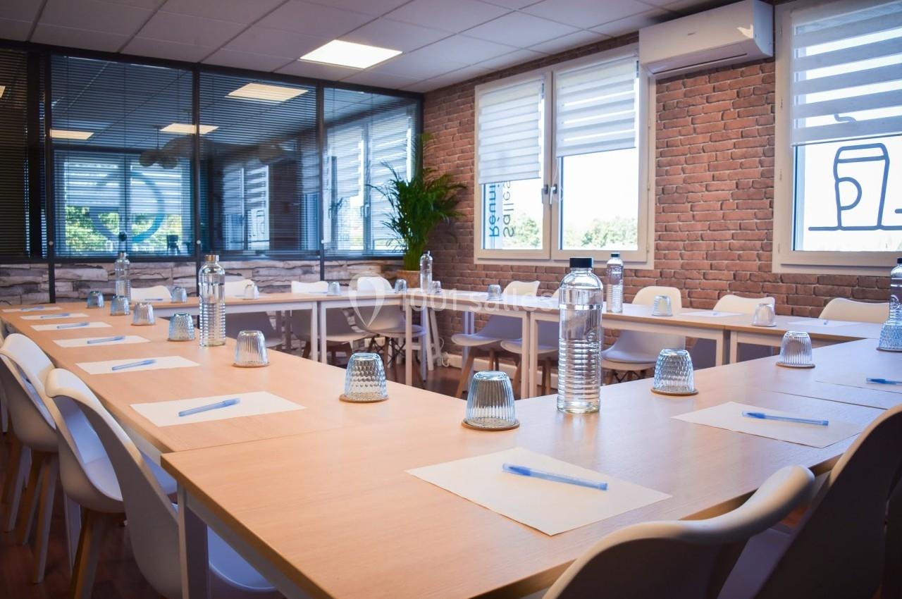 Salle de réunion lumineuse avec tables en bois disposées en U, chaises blanches, bouteilles d'eau et blocs-notes.