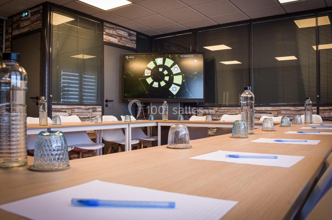 Salle de réunion moderne avec table en U, chaises blanches, bouteilles d'eau, blocs-notes, stylos et écran allumé.