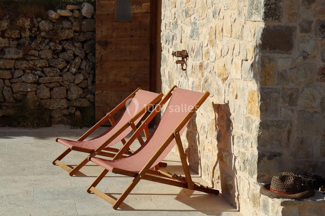 Deux chaises longues en bois et tissu rouge posées sur une terrasse en pierre, adossées à un mur en pierre.