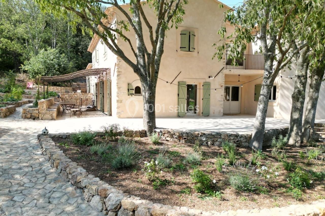 Maison en pierre claire avec volets verts, entourée d'arbres et d'un jardin aménagé avec une terrasse ombragée.