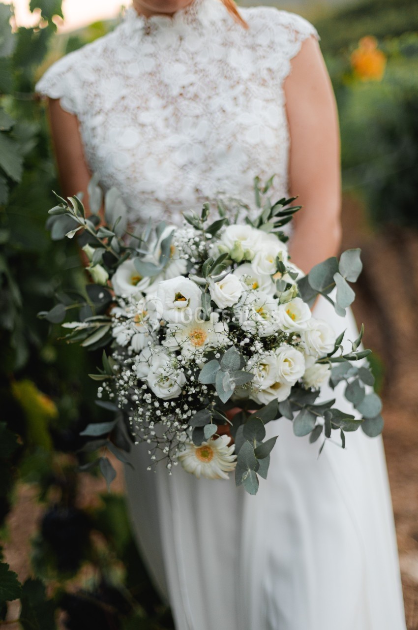 Femme en robe blanche tenant un bouquet de fleurs blanches et vertes dans un cadre naturel.
