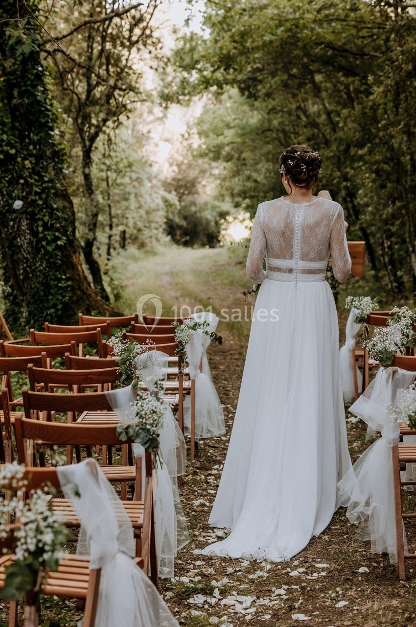 Une mariée en robe blanche marche entre des rangées de chaises décorées, dans une allée boisée.