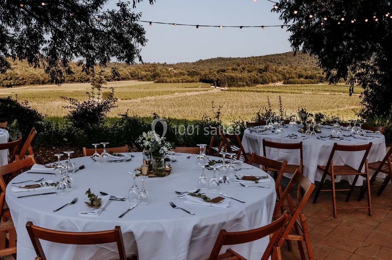 Tables rondes dressées avec nappes blanches et chaises en bois, disposées en extérieur face à un paysage de vignobles.