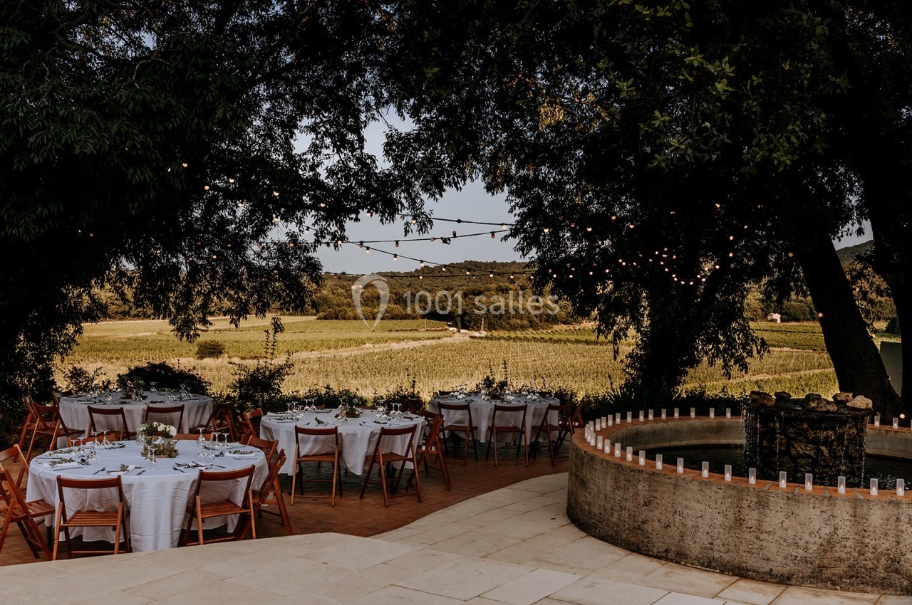 Tables rondes dressées pour un événement en plein air, entourées d'arbres et surplombant des vignes au coucher du soleil.