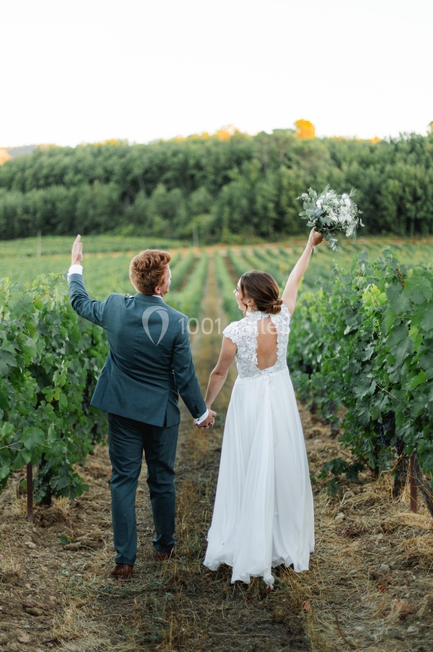 Un couple en tenue de mariage marche main dans la main dans un vignoble, la mariée levant un bouquet.