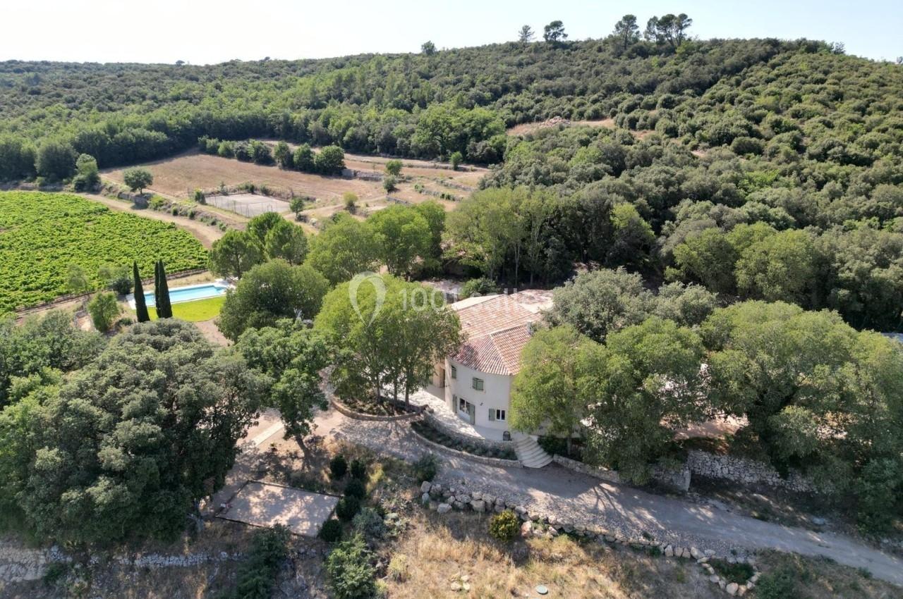 Vue aérienne d'une maison entourée d'arbres, avec une piscine et des champs dans un paysage vallonné.