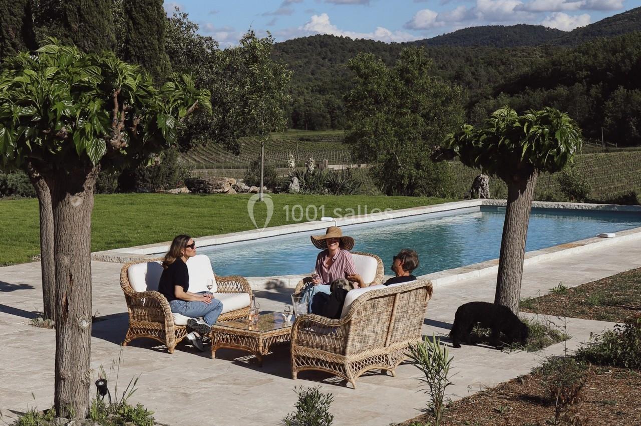 Personnes assises sur des fauteuils en rotin autour d'une table près d'une piscine, entourées de verdure et de collines.