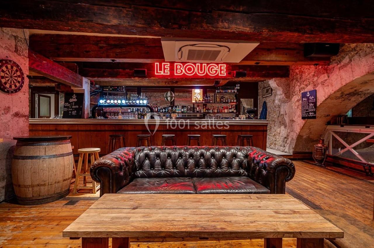 Canapé en cuir devant une table en bois dans un bar avec comptoir, enseigne lumineuse ’Le Bouge’ et décor rustique.