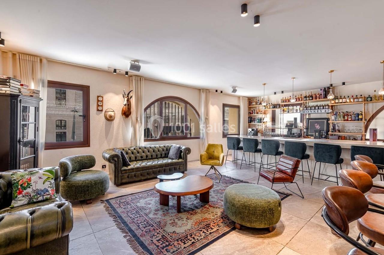 Salon spacieux avec mobilier vintage, bar en arrière-plan, grandes fenêtres et décoration éclectique.