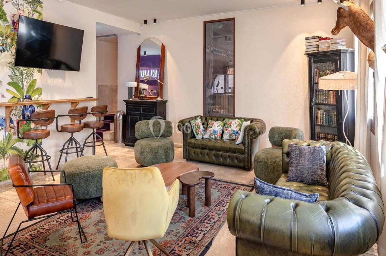 Salon lumineux avec canapés en cuir vert, fauteuils, tapis coloré, bar avec tabourets et décorations murales.