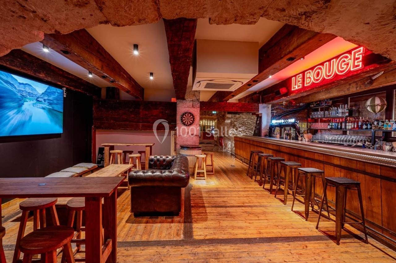 Intérieur d'un bar avec comptoir en bois, tabourets alignés, tables, canapé en cuir et enseigne lumineuse ’Le Bouge’.