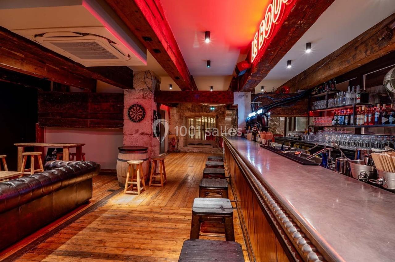 Intérieur d'un bar avec comptoir en bois, tabourets, canapé en cuir, fléchettes au mur et éclairage néon rouge.