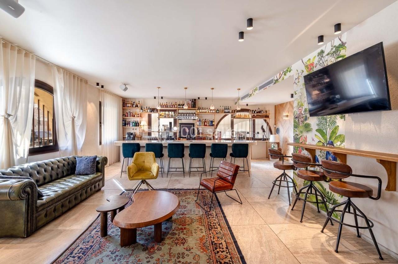 Salon moderne avec canapé en cuir, bar équipé, tabourets hauts, tapis coloré et décoration murale végétale.