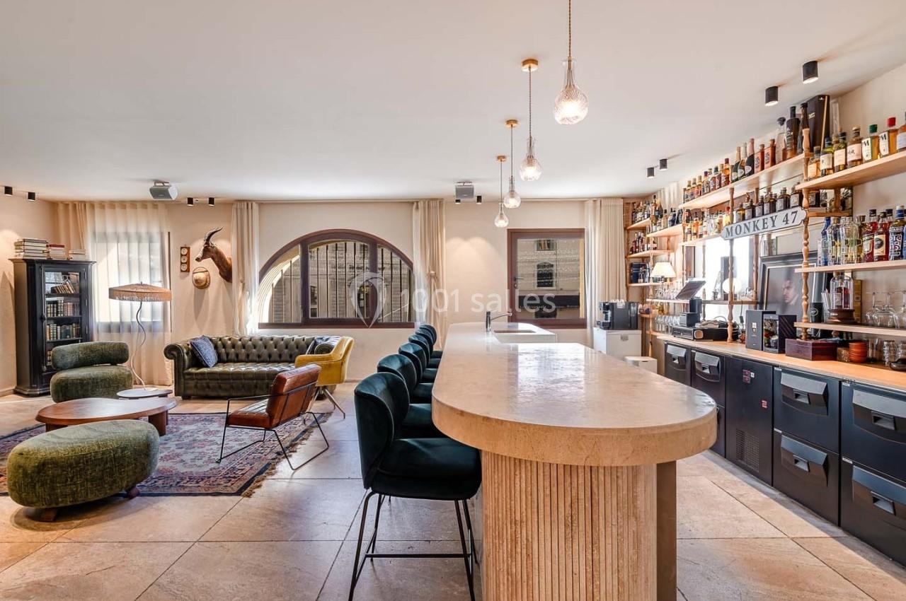 Espace intérieur lumineux avec un bar en pierre, des étagères de bouteilles, un salon et de grandes fenêtres arrondies.
