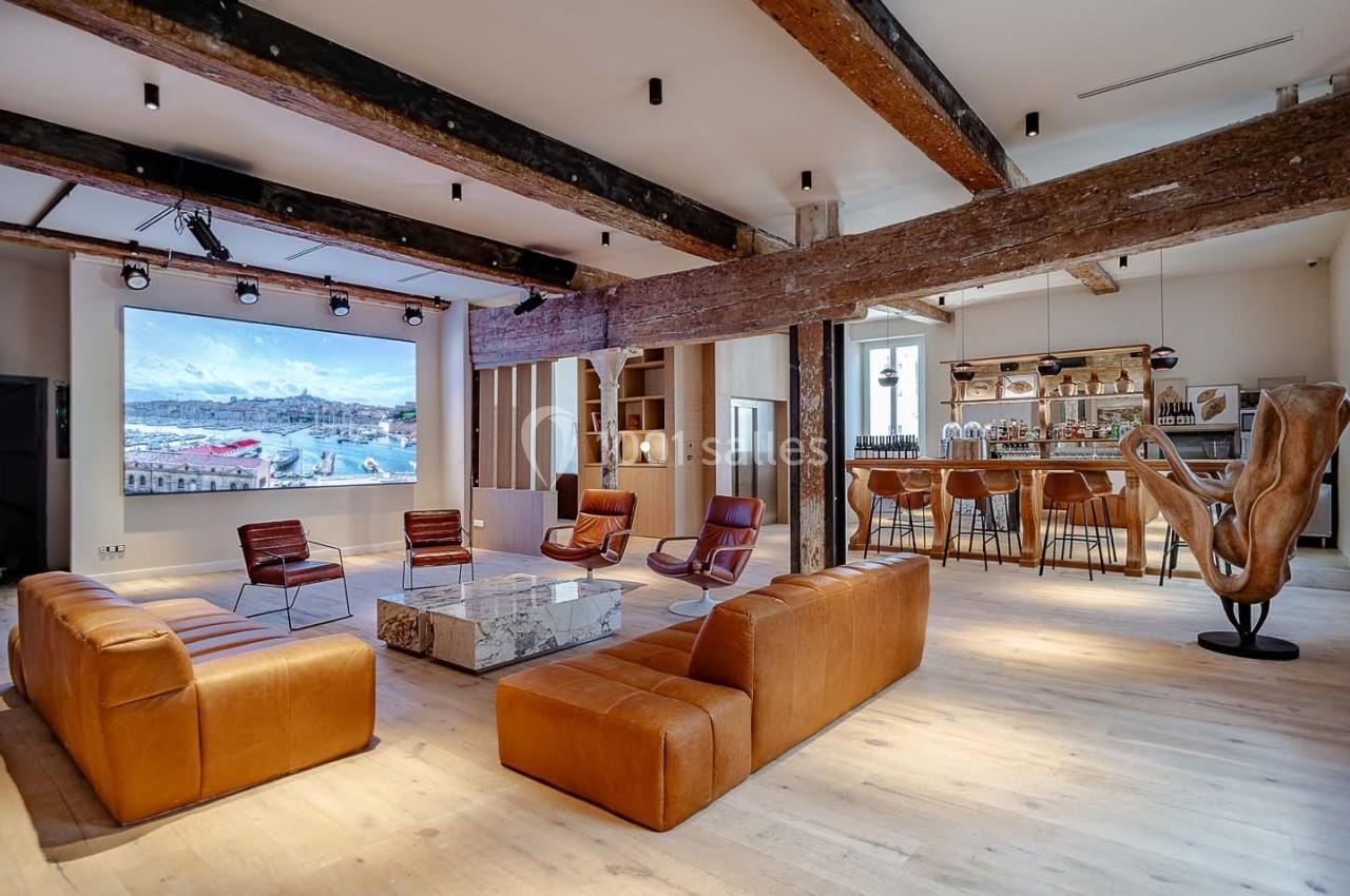Salon spacieux avec canapés en cuir, bar en bois, poutres apparentes et écran mural affichant un paysage côtier.