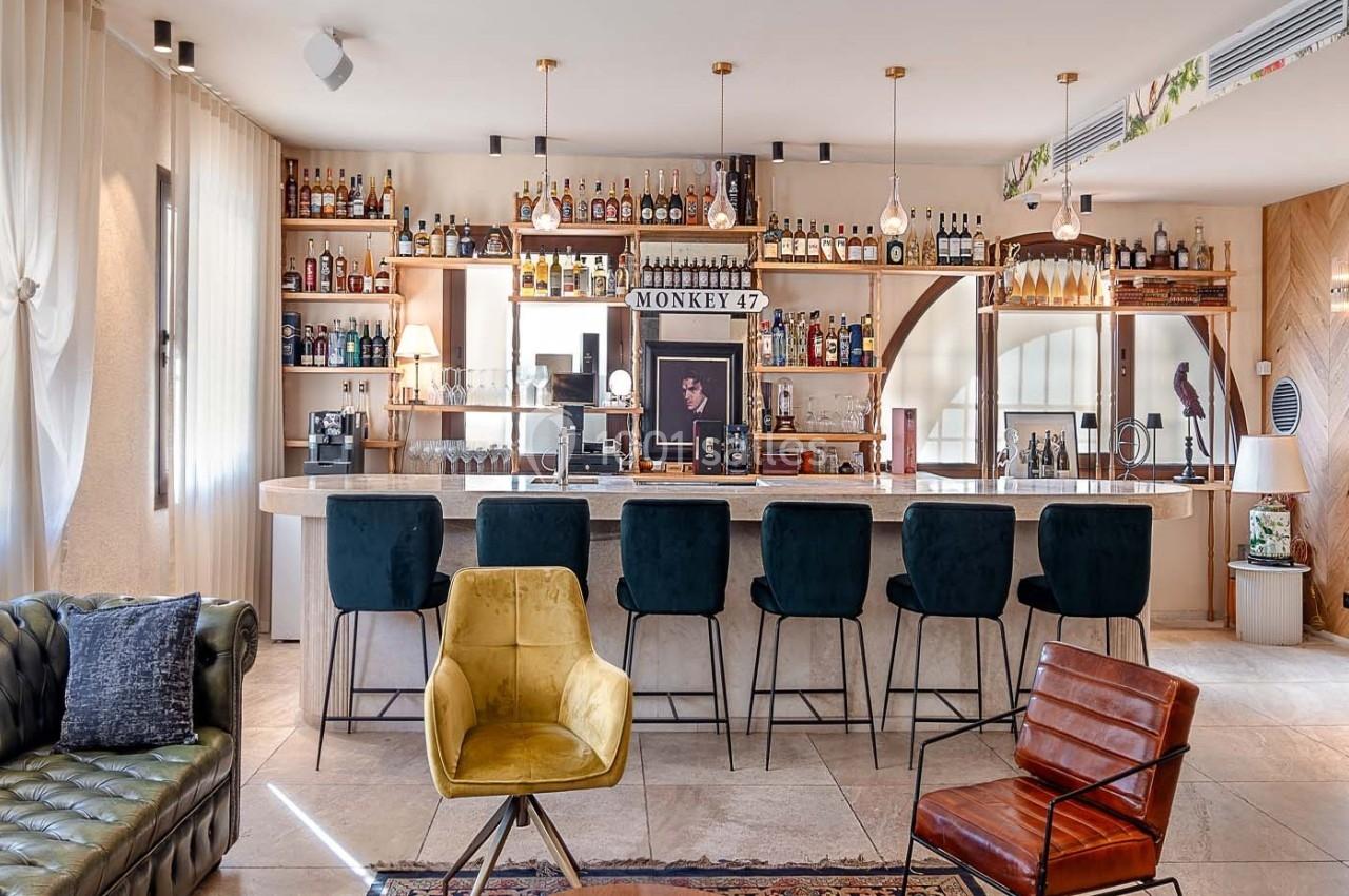 Bar moderne avec comptoir en marbre, tabourets noirs, étagères garnies de bouteilles et fauteuils design en premier plan.