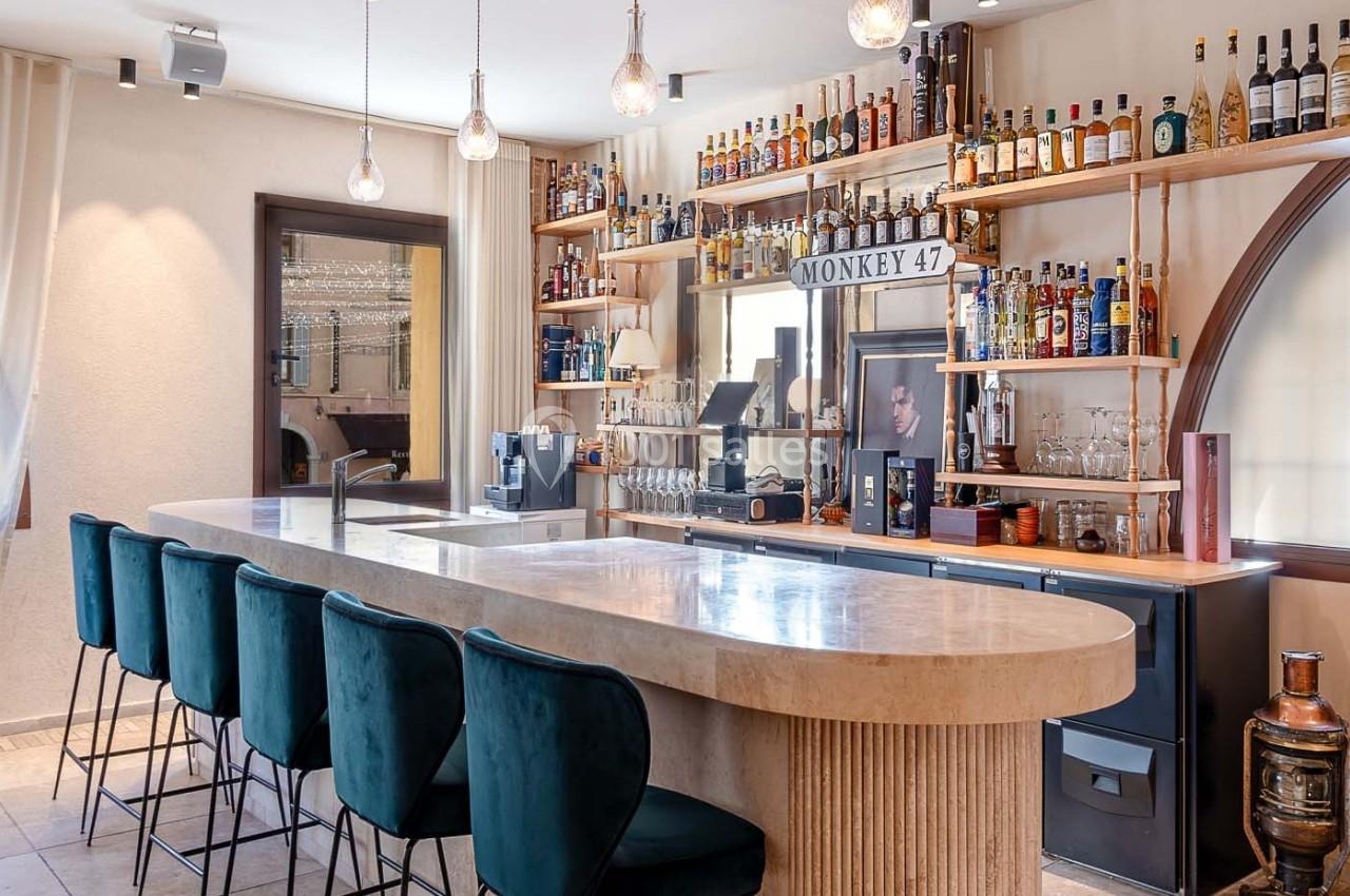 Bar moderne avec comptoir en marbre, tabourets verts, étagères remplies de bouteilles et lumière naturelle.