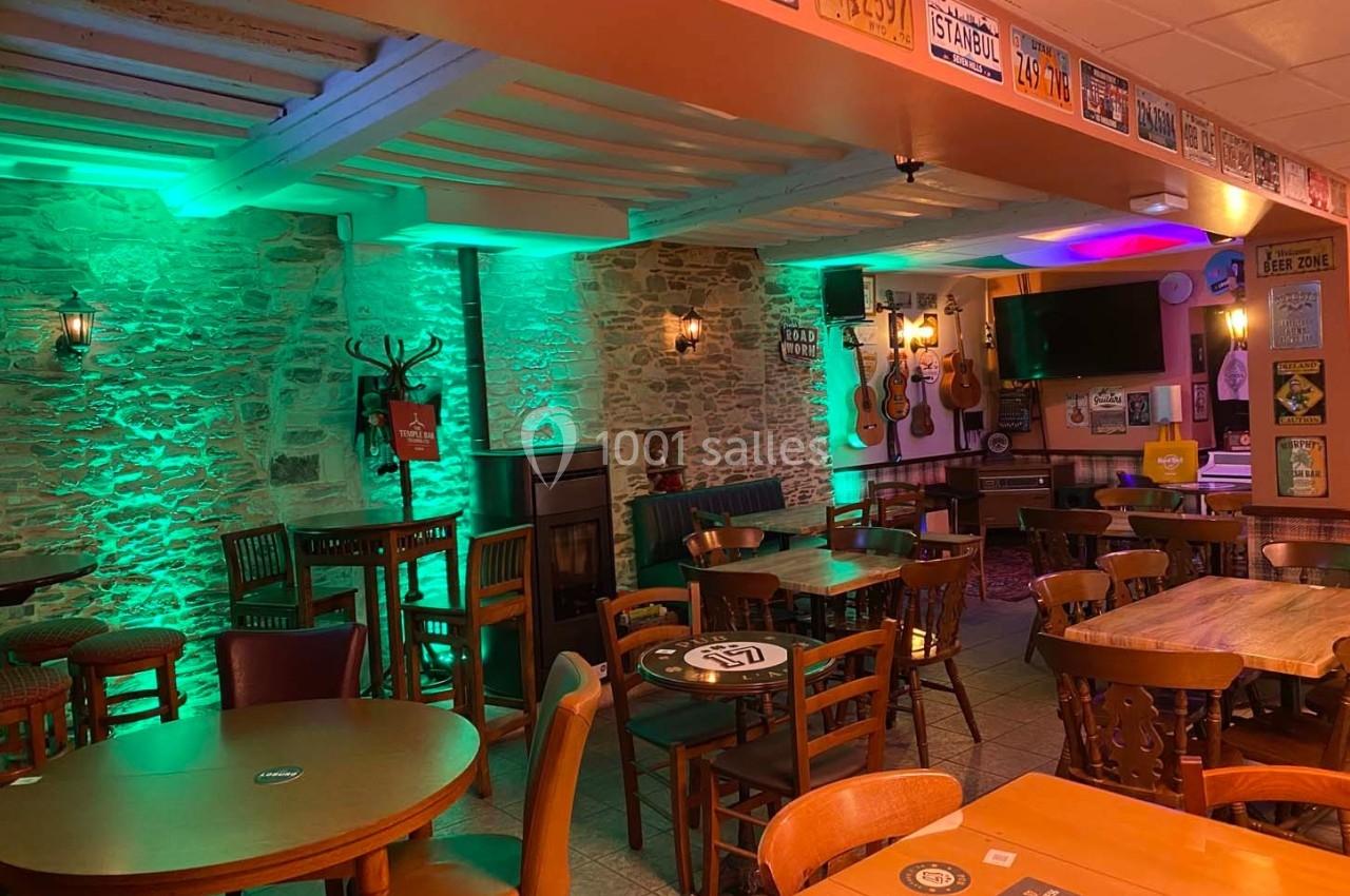 Salle de bar chaleureuse avec murs en pierre, éclairage vert et orange, tables en bois et décorations murales variées.