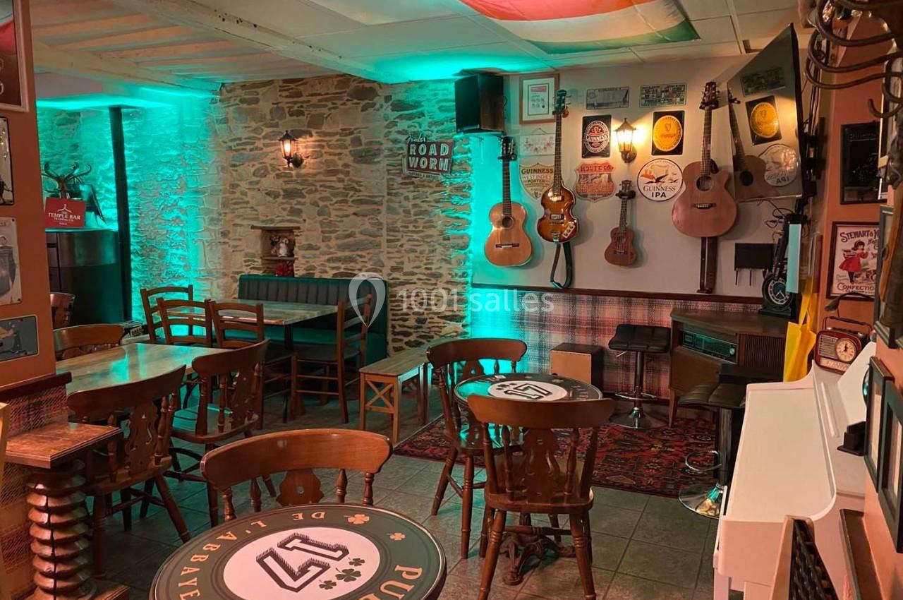 Salle de bar chaleureuse avec murs en pierre, guitares accrochées, tables en bois et éclairage vert tamisé.