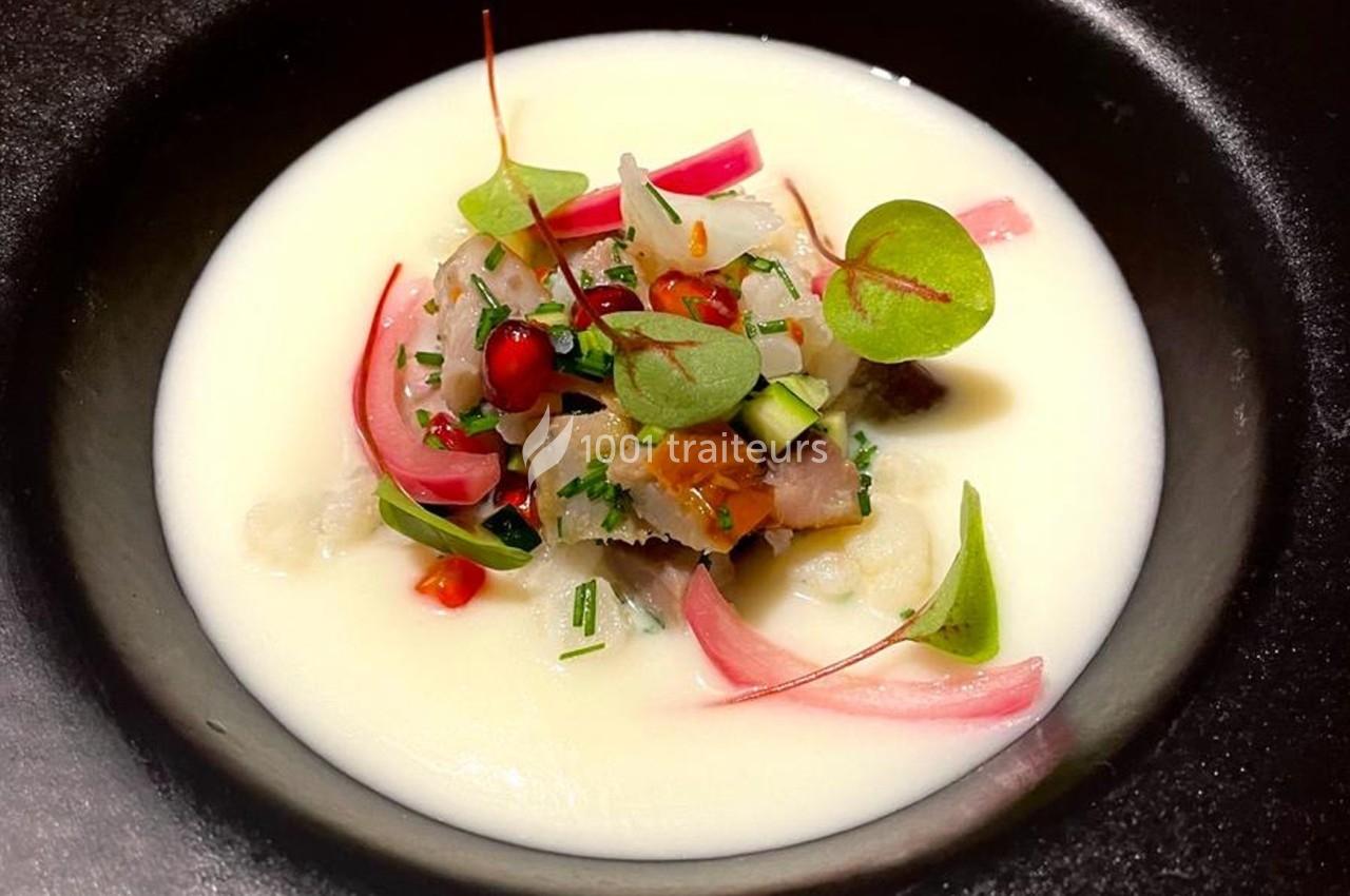 Plat gastronomique présenté dans une assiette noire, composé de poisson cru, légumes marinés et herbes sur une sauce blanche.