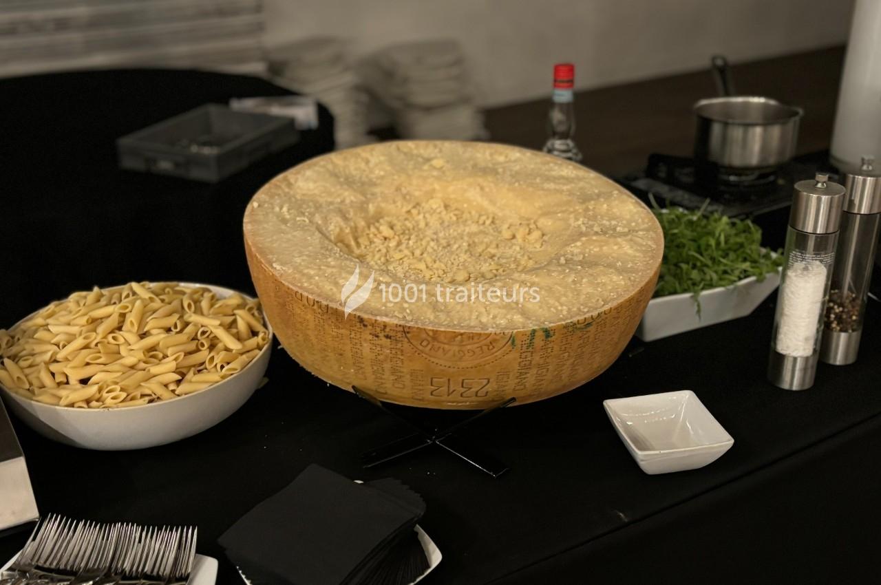 Grande meule de fromage creusée entourée de pâtes, roquette, condiments et ustensiles sur une table noire.