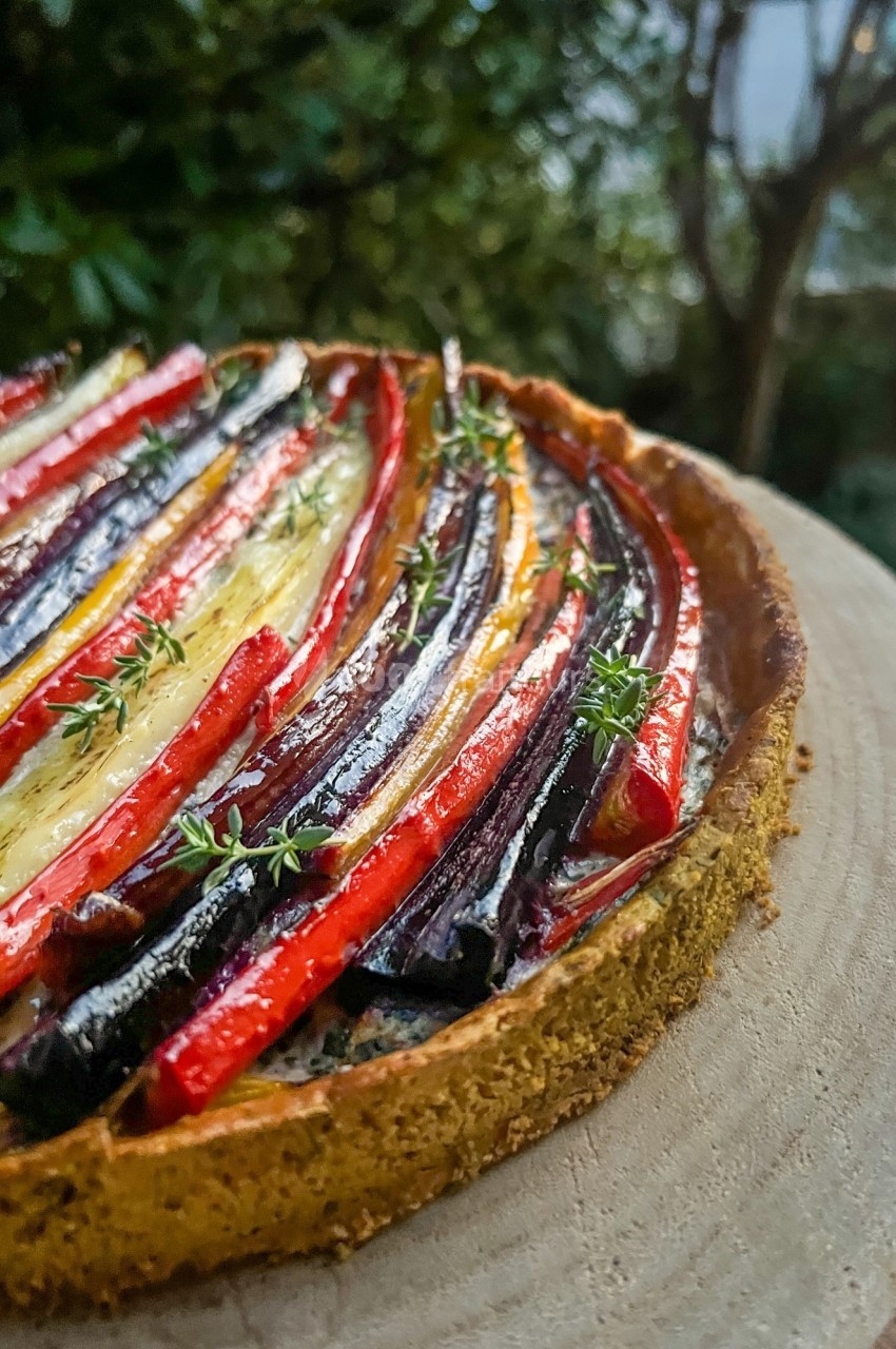 Tarte salée garnie de légumes colorés disposés en rangées sur une pâte dorée, posée sur une planche en bois.