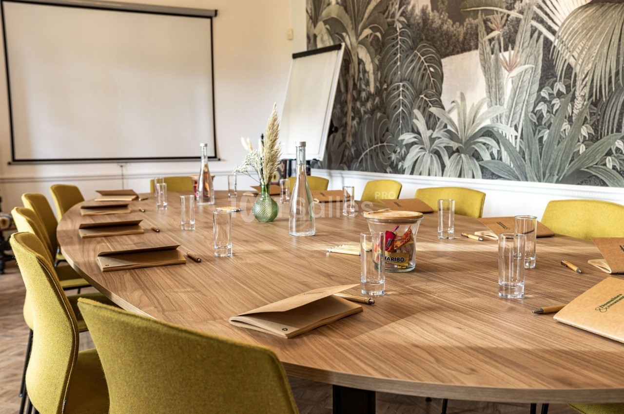 Salle de réunion avec une grande table ovale en bois, chaises jaunes, fournitures de bureau et un écran de projection.