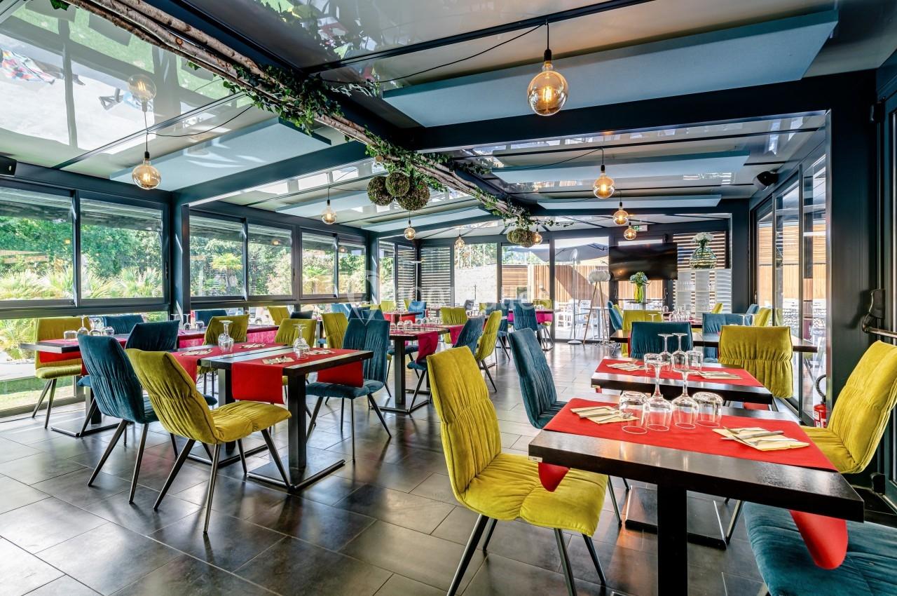 Salle de restaurant lumineuse avec grandes baies vitrées, tables dressées et chaises colorées.