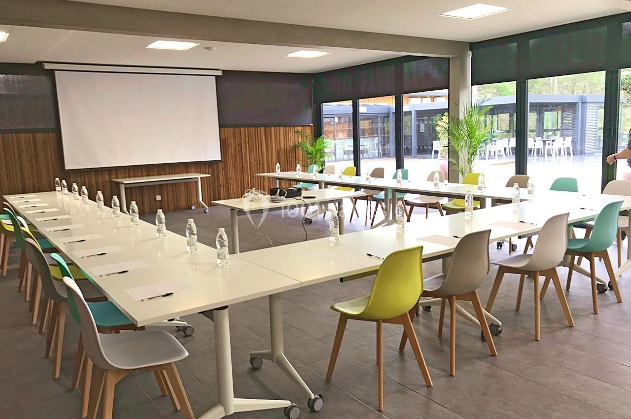 Salle de réunion lumineuse avec tables disposées en U, chaises colorées, écran de projection et grandes baies vitrées.