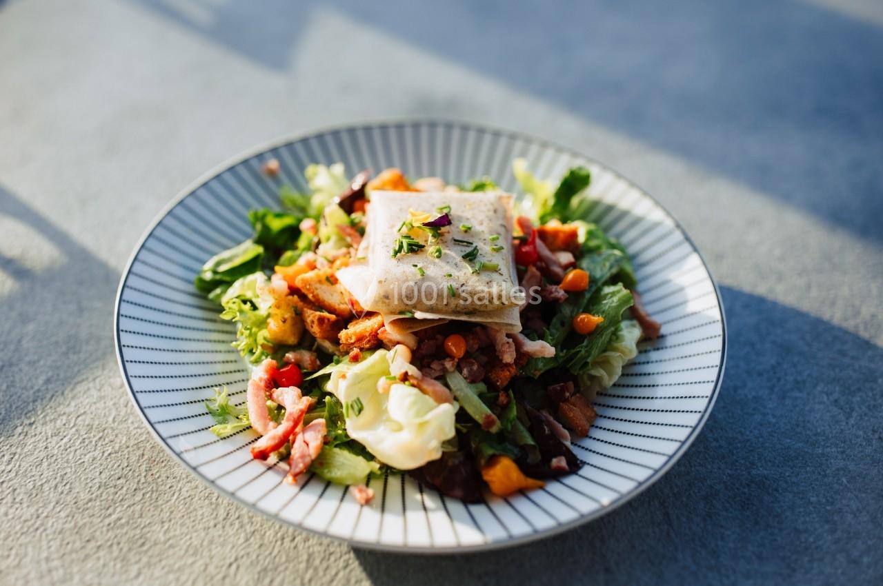 Assiette de salade composée avec légumes, lardons, croûtons et un carré de fromage fondu, servie sur une table grise.