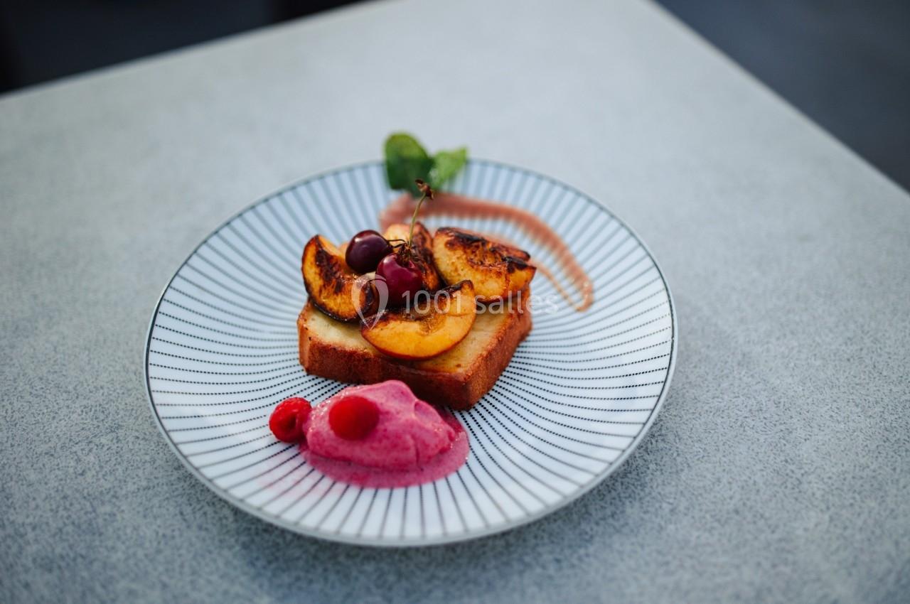 Tranche de gâteau garnie de pêches grillées, cerises, framboises et crème rose, servie sur une assiette blanche rayée.