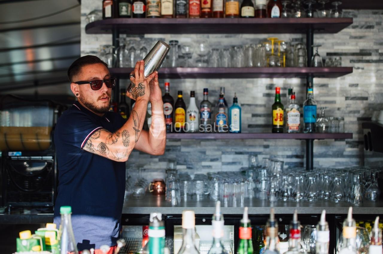 Un barman tatoué mélange un cocktail dans un shaker, devant un bar garni de bouteilles et de verres.