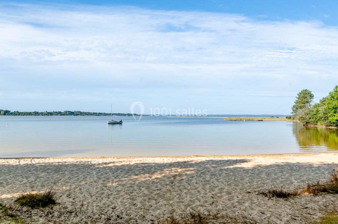Vue d'une plage de sable avec un voilier ancré sur un lac calme, entouré de végétation et d'un ciel dégagé.